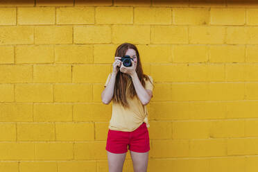 Young woman photographing through camera while standing against yellow wall - MGRF00027