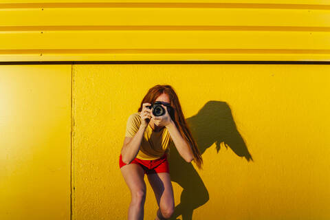Woman photographing through camera while standing against yellow wall stock photo