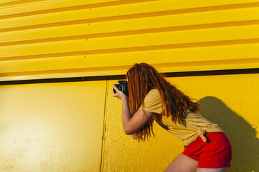Rothaarige Frau, die durch die Kamera fotografiert, während sie vor einem gelben Hintergrund steht - MGRF00029