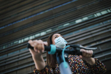 Mature woman in protective face mask with electric scooter against building during coronavirus crisis - GRCF00433