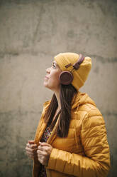 Thoughtful woman in warm clothing listening music through headphones while standing by wall - GRCF00442