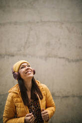 Happy mature woman in warm clothing looking up while listening music through headphones against wall - GRCF00443