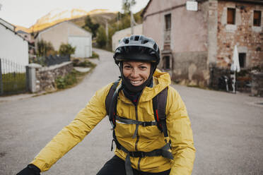 Smiling mid adult woman in warm clothing riding mountain bike on road while traveling to Somiedo Natural Park, Spain - DMGF00239