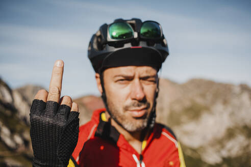 Mittlerer erwachsener Mann zeigt den Mittelfinger, während er gegen einen Berg im Naturpark Somiedo, Spanien, steht - DMGF00248