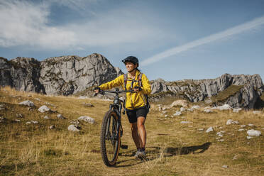 Mittlere erwachsene Frau mit Fahrrad vor einem Berg im Naturpark Somiedo, Spanien - DMGF00260