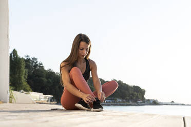 Sportlerin beim Binden von Schnürsenkeln auf der Promenade am Meer bei Sonnenaufgang - AFVF07395