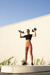 Sportswoman flexing muscle with dumbbell standing on pedestal against wall during sunrise - AFVF07407