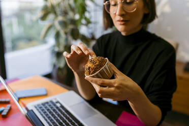 Geschäftsfrau hält Kuchen, während sie zu Hause am Laptop sitzt - VABF03811