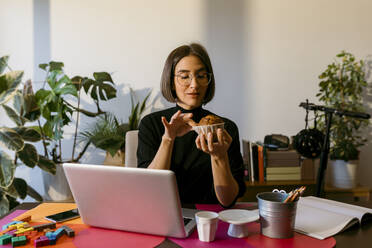 Mid adult businesswoman holding cake while sitting by laptop at home - VABF03812