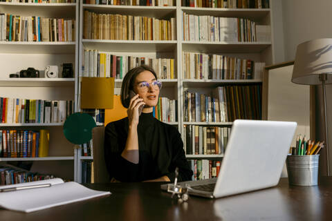 Businesswoman talking on smart phone looking away sitting by laptop against bookshelf at home stock photo