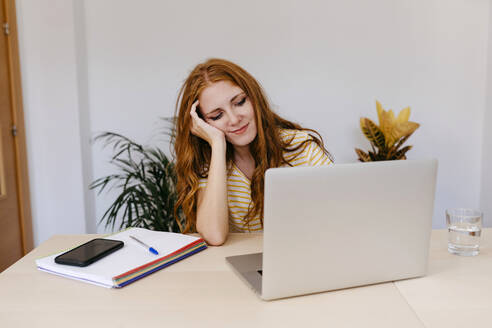 Junge Frau mit Hand in den Haaren, die einen Laptop benutzt, während sie zu Hause sitzt - TCEF01262