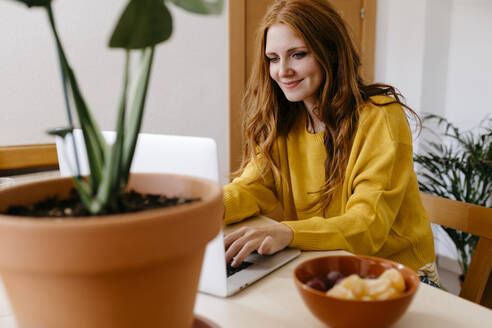 Lächelnde Frau, die einen Laptop benutzt, während sie zu Hause sitzt - TCEF01265