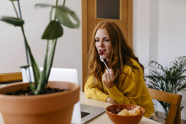 Junge Frau isst Obst, während sie zu Hause einen Laptop benutzt - TCEF01266