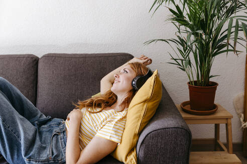 Junge Frau hört Musik über Kopfhörer, während sie zu Hause auf dem Sofa liegt - TCEF01271