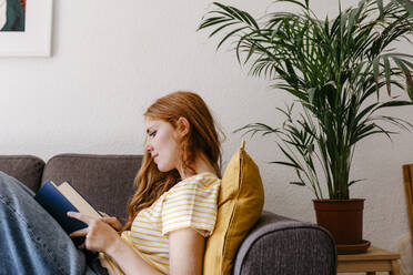 Junge Frau liest ein Buch, während sie zu Hause auf dem Sofa liegt - TCEF01273