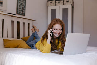 Lächelnde Frau, die auf der Vorderseite liegt und mit ihrem Smartphone im Schlafzimmer telefoniert - TCEF01279