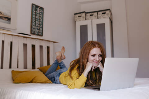 Lächelnde Frau auf der Vorderseite liegend mit der Hand am Kinn, die zu Hause einen Laptop benutzt, lizenzfreies Stockfoto