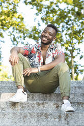 Smiling young man in casuals sitting on steps against trees - PNAF00028