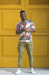 Happy young man with hands clasped standing against closed yellow doors - PNAF00033