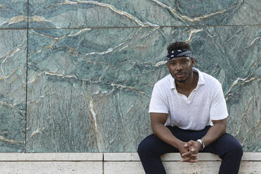 Young man wearing headscarf while sitting against wall - PNAF00041