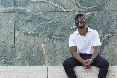 Cheerful young man in headscarf sitting against patterned wall - PNAF00042