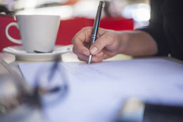 Junge Frau mit Kaffeetasse schreibt in Papier, während sie in einem Straßencafé sitzt - AJOF00493