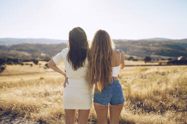 Weibliche Freunde betrachten die Aussicht, während sie auf einem grasbewachsenen Feld gegen den Himmel an einem sonnigen Tag stehen - RSGF00395
