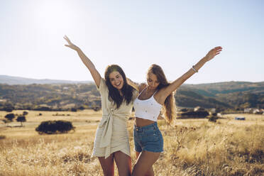 Cheerful young female friends standing with hands raised on grassy field against sky - RSGF00397