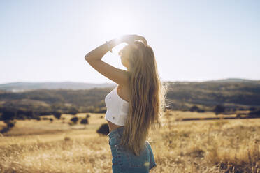 Young woman with hand in hair standing on field against sky during sunny day - RSGF00400