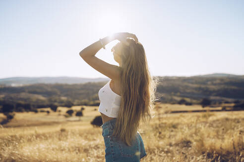 Junge Frau mit Hand in den Haaren steht auf einem Feld gegen den Himmel an einem sonnigen Tag - RSGF00400
