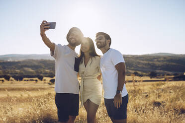 Lächelnde männliche und weibliche Freunde, die ein Selfie mit ihrem Smartphone machen, während sie auf einem Feld im Sommer stehen - RSGF00401