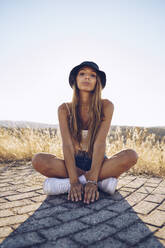 Young woman sitting on footpath against sky on sunny day - RSGF00403