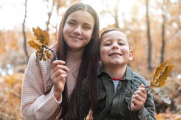 Brother and sister holding dry leaves while standing in Cannock Chase park during autumn - WPEF03514