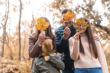 Familie, die ihr Gesicht mit trockenen Blättern bedeckt, im Herbst im Park stehend - WPEF03515