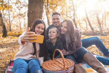 Glückliche Familie, die ein Selfie mit ihrem Smartphone macht, während sie im Herbst im Park sitzt - WPEF03524