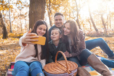 Happy family taking selfie on smart phone while sitting in park during autumn - WPEF03524