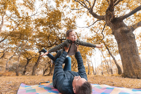 Junge, der sich vorstellt, dass er fliegt, gehalten von seinem Vater, auf einer Picknickdecke im Park im Herbst - WPEF03539