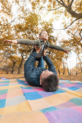 Junge, der sich vorstellt, dass er fliegt, gehalten von seinem Vater, der auf einer Picknickdecke im Park liegt - WPEF03540