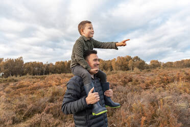 Vater, der seinen Sohn auf der Schulter trägt, steht im Herbst im Park von Cannock Chase vor einem bewölkten Himmel - WPEF03573