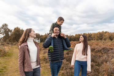 Father carrying son on shoulder while mother and sister standing beside in park during autumn - WPEF03576