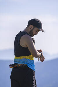 Mann mit Mütze prüft die Zeit, während er vor einem Berg und einem klaren Himmel steht - SNF00759