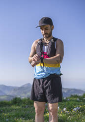 Mittlerer erwachsener Mann mit Mütze und Flaschengurt, der auf einem Berg stehend die Zeit auf der Uhr überprüft - SNF00760