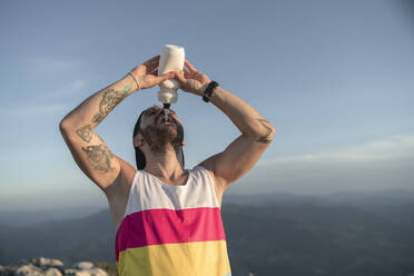 Athlete drinking water while standing on mountain against clear sky - SNF00779