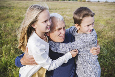 Grandfather looking away while embracing cute grandchildren on field - EYAF01382