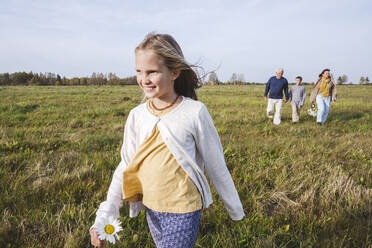 Lächelndes Mädchen, das auf einem Feld läuft, während die Familie im Hintergrund zu sehen ist - EYAF01388