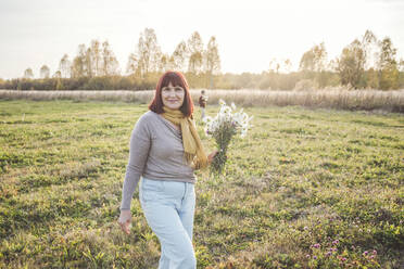 Senior woman walking with bouquet of chamomile flowers on field during sunny day - EYAF01409
