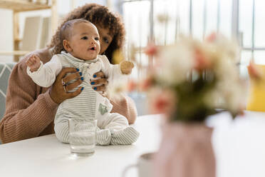 Mother looking at cute daughter sitting on table in living room - GIOF09512