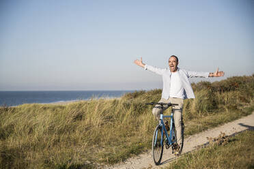 Glücklicher reifer Mann mit ausgestreckten Armen beim Fahrradfahren am Strand gegen den klaren Himmel - UUF21973