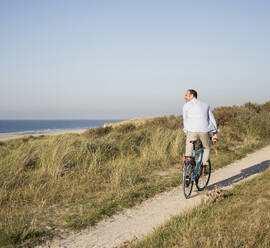 Fröhlicher reifer Mann fährt Fahrrad am Strand gegen den klaren Himmel - UUF21976