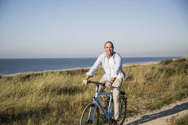 Smiling mature man riding bicycle at beach against clear sky - UUF21978